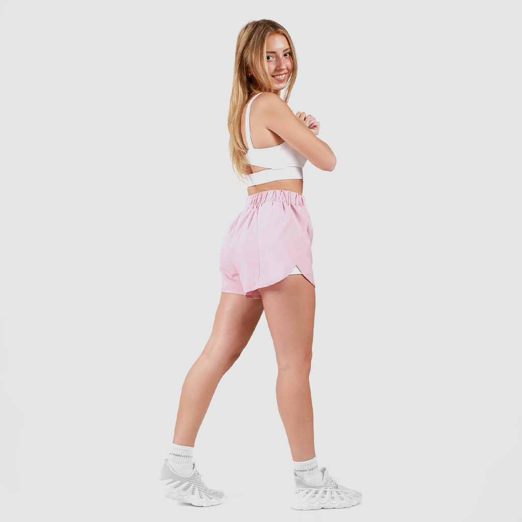 a fit woman stand a quarter pose 
 and look at the camera ,she wearing sport white bra with  short pink ,and she wearing shoes white ,plus the  background is light grey  