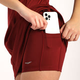 A person wearing red athletic shorts with a built-in pocket on the side, placing a white smartphone  (iPhone) into the pocket. The shorts have a small logo on the lower part of the leg.