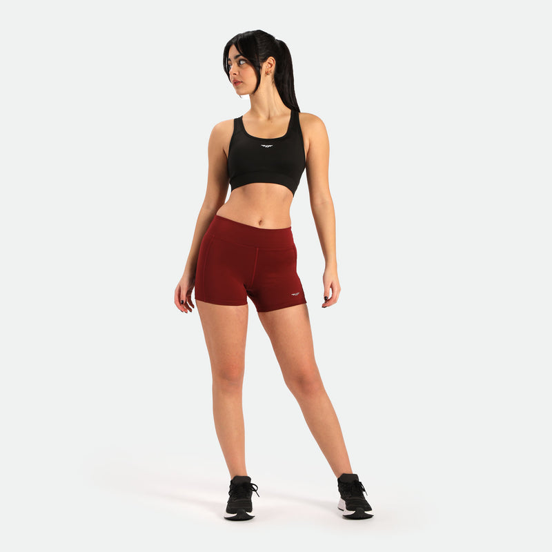 a fit woman stand with pose and look at right ,she wearing  black sports bra and short red with logo , and she wearing shoes black ,plus the background is light grey 