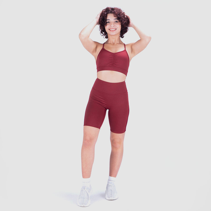 a fit woman  look at the camera with pose and smile ,she wearing matching sport bra and seamless short red with socks and shoes white ,plus the background is light grey 