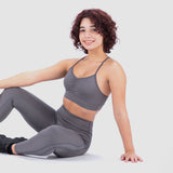 A woman with curly hair wearing a matching gray sports bra and leggings is sitting on the floor in a relaxed pose, smiling at the camera against a plain white background