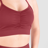 A close-up of a woman's torso wearing a burgundy sports bra with rushed detailing at the center and matching high-waisted leggings. The background is plain light gray