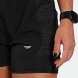 Person wearing black athletic shorts with a logo, holding a phone, on a neutral background