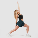A woman wearing a black sports bra, dark teal athletic shorts, and white sneakers is performing a yoga pose. She is in a lunge position with her front leg bent and back leg straight, reaching one arm up toward the sky and the other arm down toward her back leg. Her gaze follows her raised hand. The background is plain white