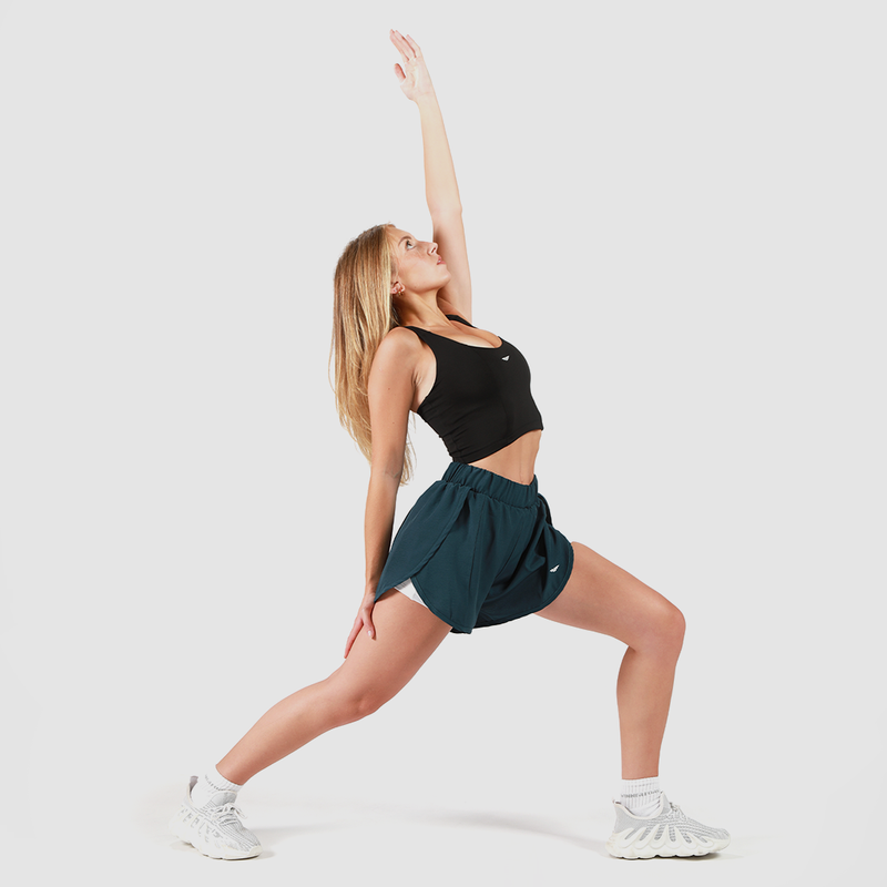 A woman wearing a black sports bra, dark teal athletic shorts, and white sneakers is performing a yoga pose. She is in a lunge position with her front leg bent and back leg straight, reaching one arm up toward the sky and the other arm down toward her back leg. Her gaze follows her raised hand. The background is plain white