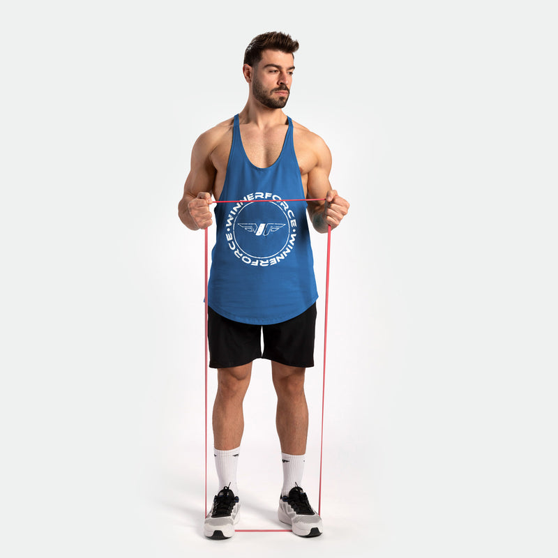 A man in a blue tank top and black shorts exercises with a red resistance band. He stands confidently, wearing white sneakers, on a plain background.