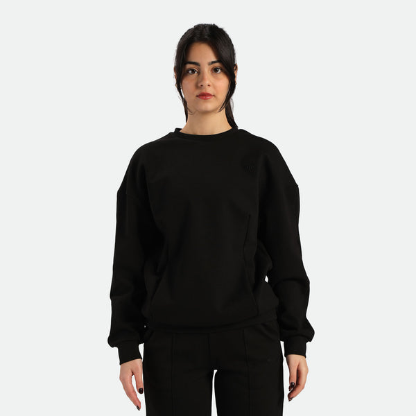 A young woman stands facing forward, wearing a black sweatshirt and black pants, against a plain white background.