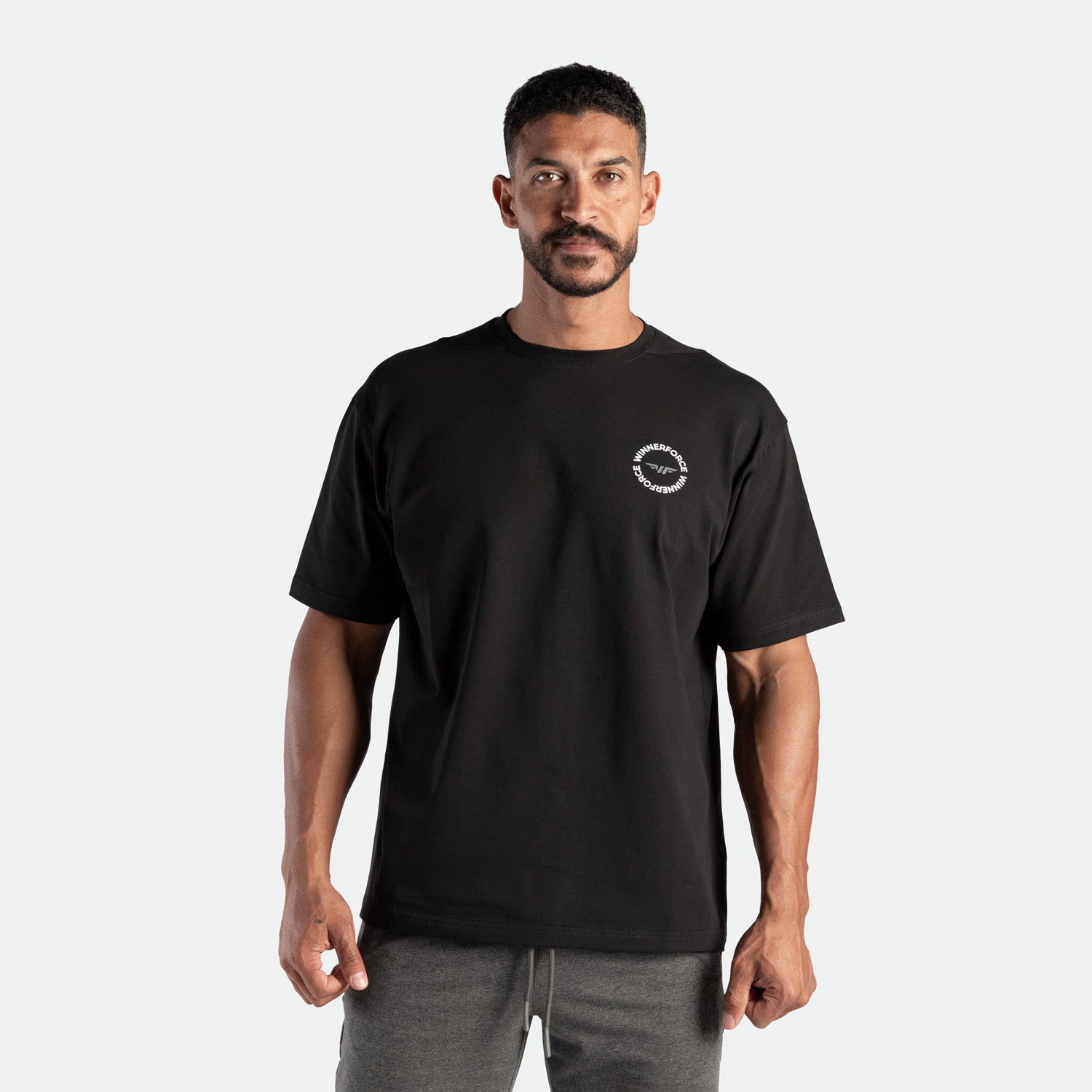 a fit man look at the camera and wearing t-shirt black with logo, plus the pants grey 