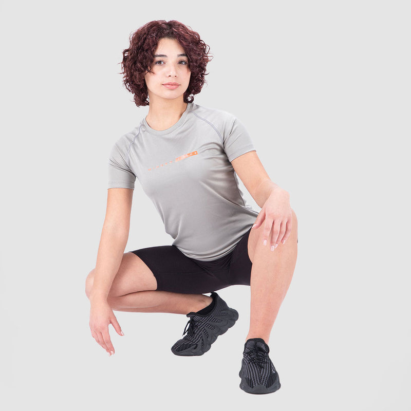 Woman with curly hair squats against a light gray background, wearing a gray T-shirt, black shorts, and black athletic shoes, projecting confidence.