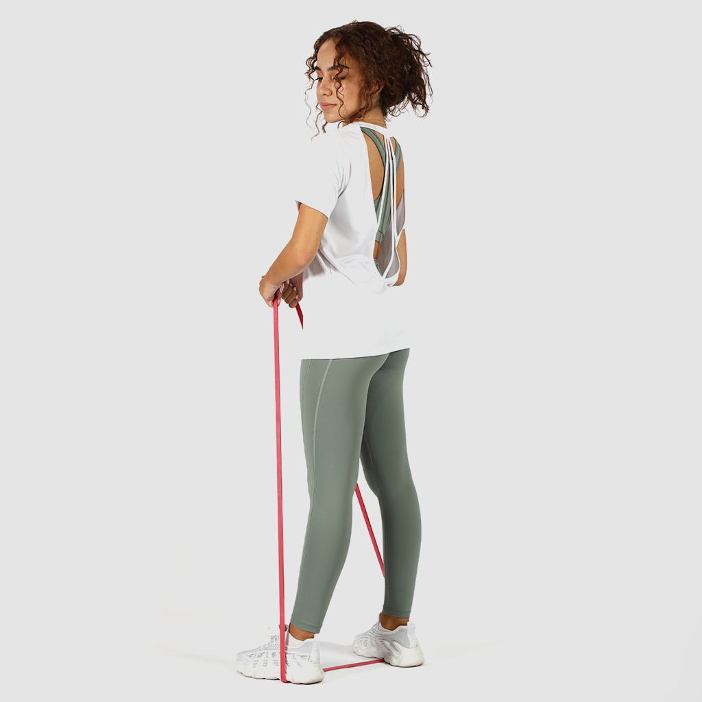 A woman with curly hair tied back is exercising with a red resistance band. She is wearing a white open-back T-shirt with strap details and a sage green sports bra underneath. She is also dressed in matching sage green leggings and white athletic shoes