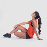 Woman in an orange tank top and black shorts sits on the floor, leaned back on her hand, with her head raised, appearing relaxed and content.