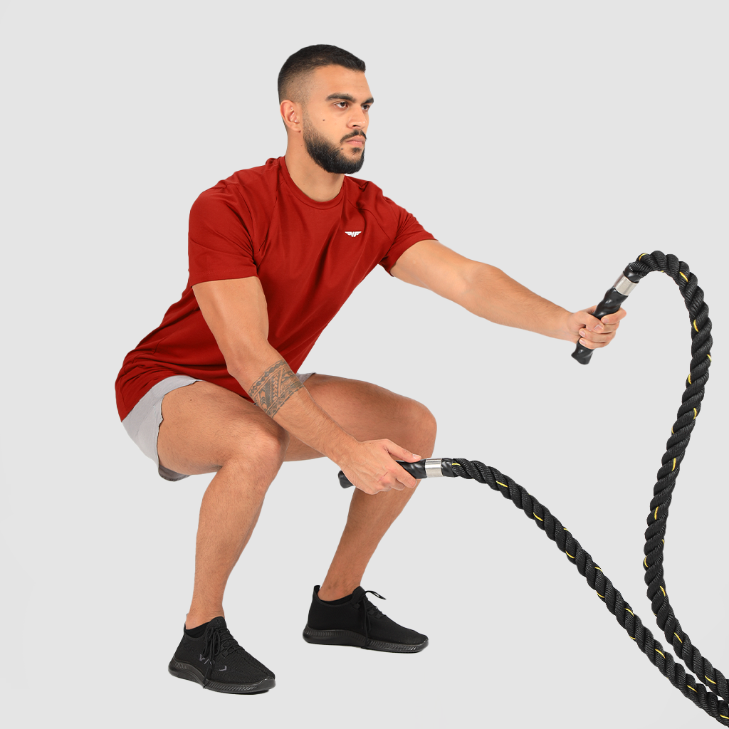 Man wearing a red athletic t-shirt and light gray workout shorts performing battle rope exercise in a squat position