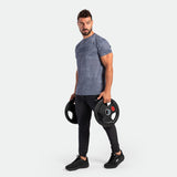 A muscular man in a gray camo shirt and black leggings holds two weight plates, conveying strength and focus, against a plain white background.