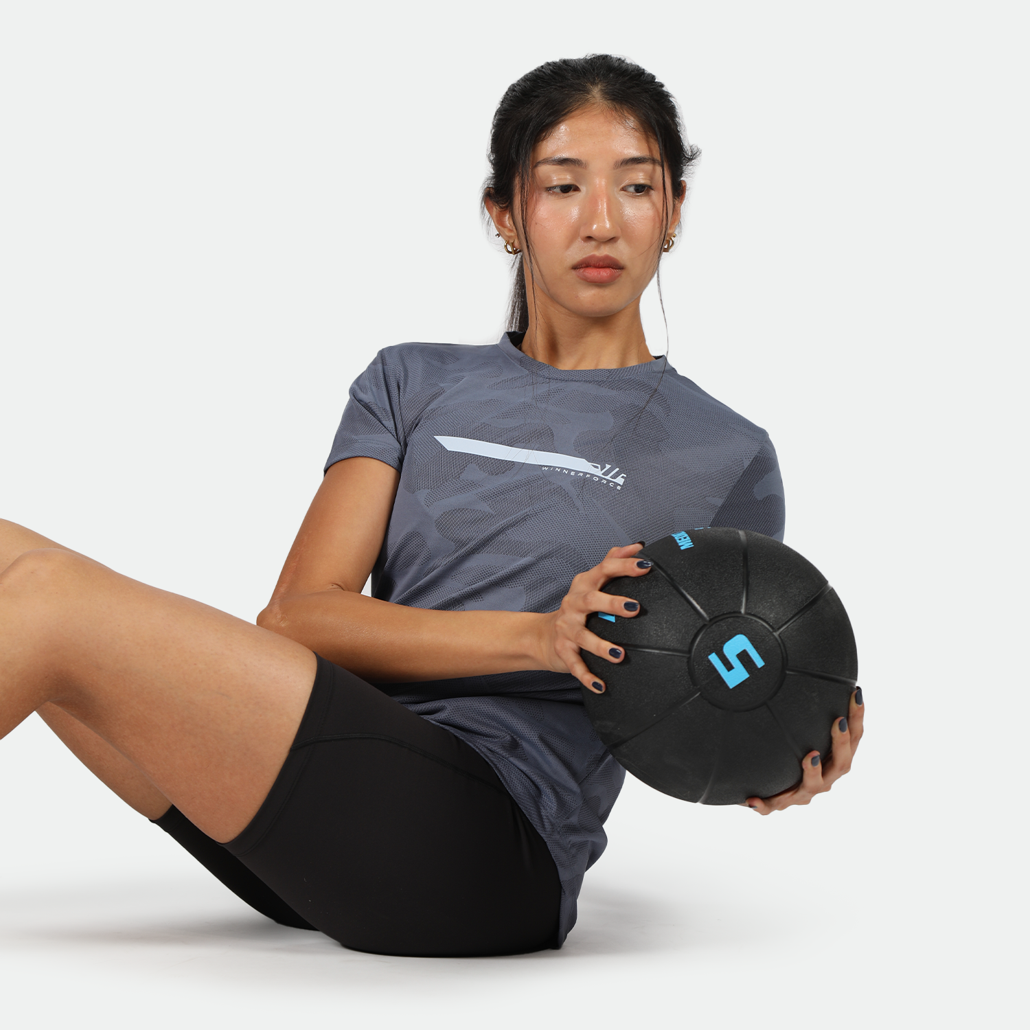 A woman is performing a Russian twist exercise while holding a black 5-pound medicine ball. She is wearing a blue camo-patterned athletic shirt and black workout shorts, seated on the floor with her legs bent and torso slightly twisted.