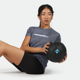 A woman is performing a Russian twist exercise while holding a black 5-pound medicine ball. She is wearing a blue camo-patterned athletic shirt and black workout shorts, seated on the floor with her legs bent and torso slightly twisted.