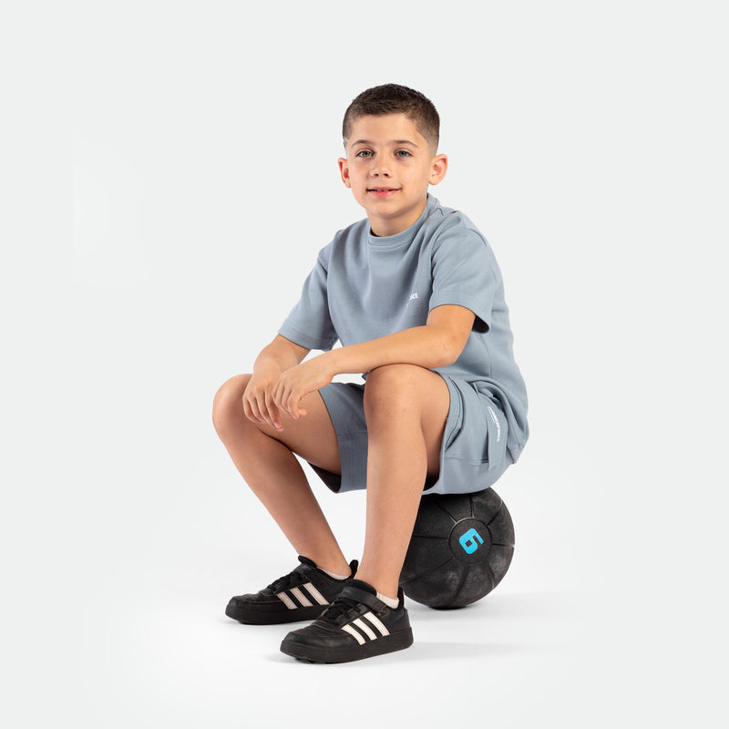 Young boy in a light blue outfit sits on a black medicine ball with a blue "6," against a white background. He wears black Adidas sneakers and smiles softly.