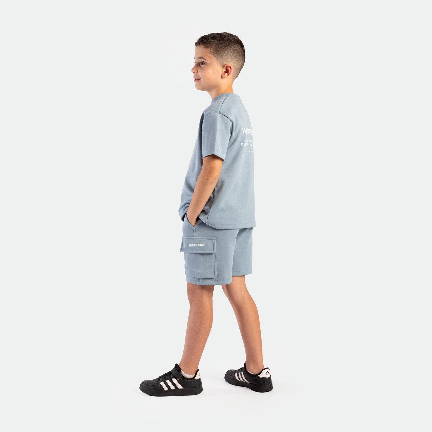 a young  boy stand at right and wearing matching set dusty blue ,t-shirt  with logo and short with pocket , plus the shoes 