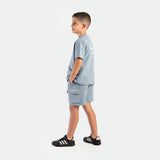 a young  boy stand at right and wearing matching set dusty blue ,t-shirt  with logo and short with pocket , plus the shoes 