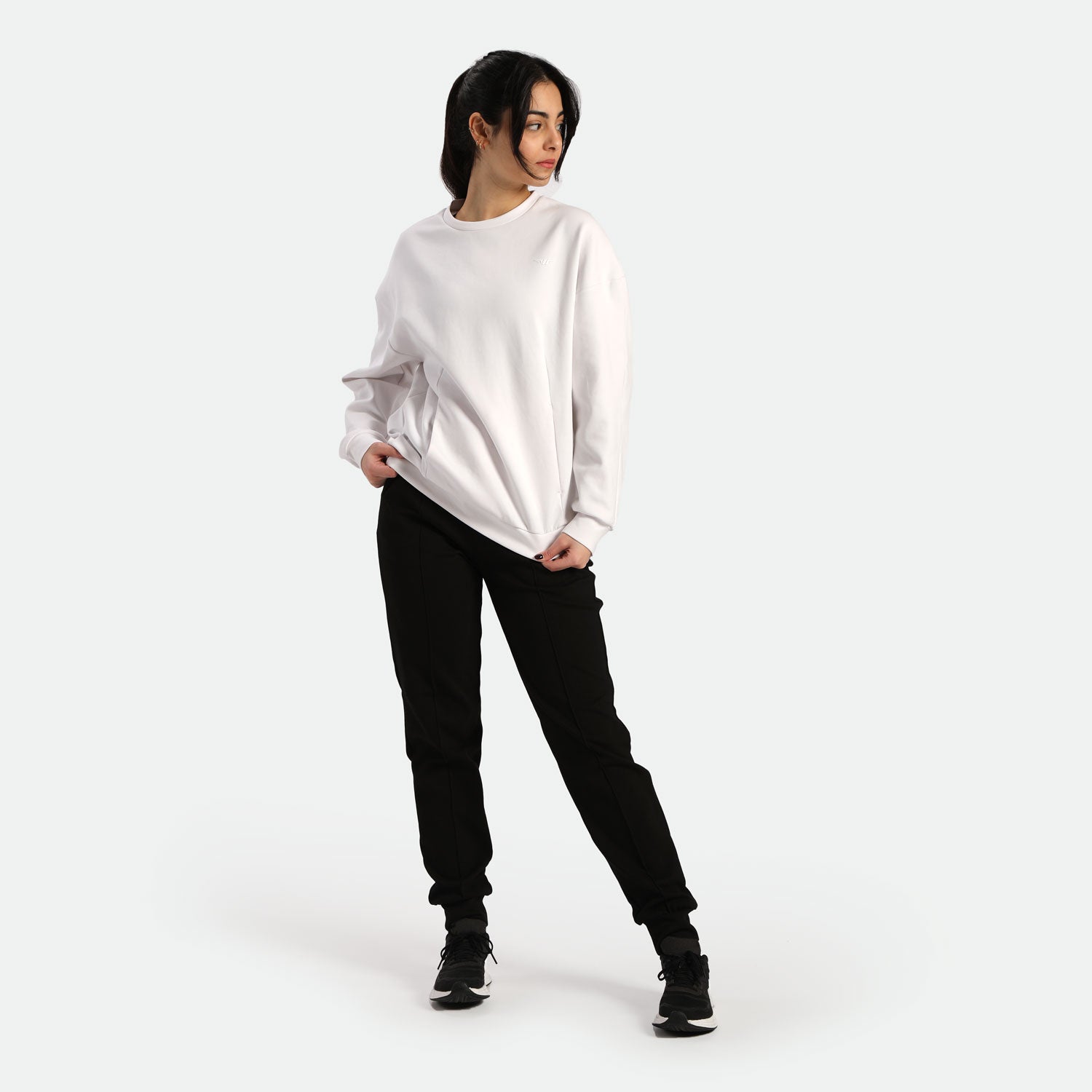 A young woman stands casually in a white sweatshirt and black joggers, posing confidently against a plain light background.
