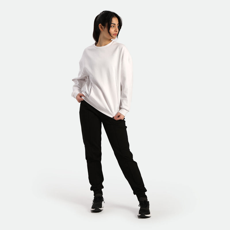 A young woman stands casually in a white sweatshirt and black joggers, posing confidently against a plain light background.