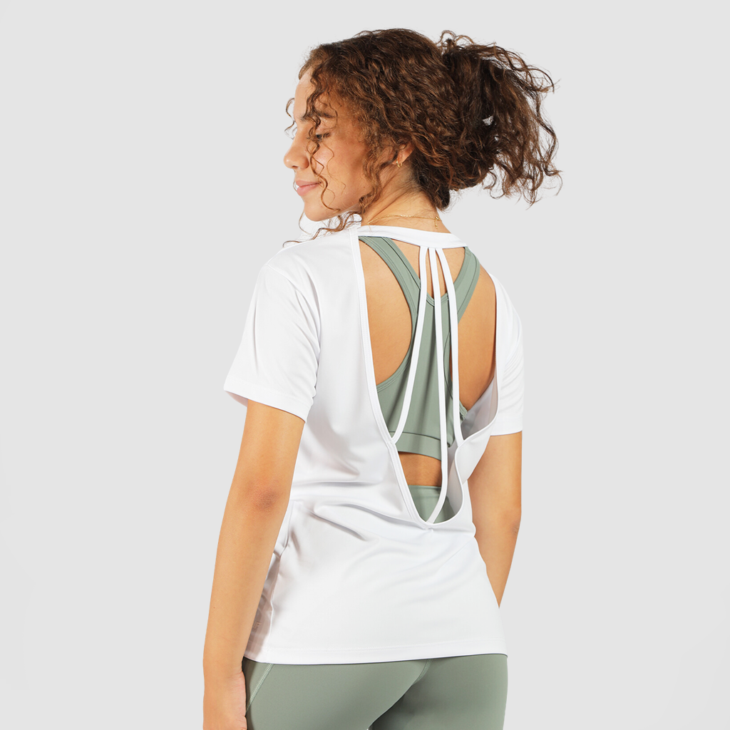 A woman with curly hair tied in a ponytail is wearing a white short-sleeved T-shirt with an open back design that features two straps tying at the top. Underneath, she is wearing a sage green sports bra. She is also wearing matching sage green leggings