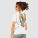 A woman with curly hair tied in a ponytail is wearing a white short-sleeved T-shirt with an open back design that features two straps tying at the top. Underneath, she is wearing a sage green sports bra. She is also wearing matching sage green leggings