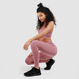 A woman with curly hair wearing a matching pink sports bra and leggings, crouching down with one arm resting on her knee, dressed in black athletic shoes against a plain gray background