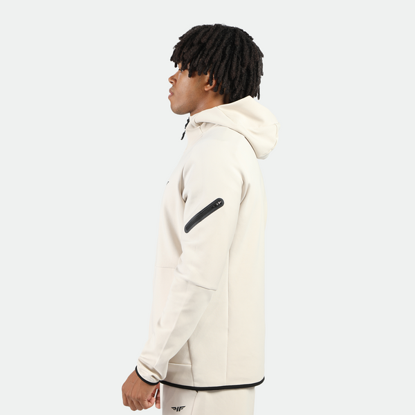 a fit man stand at right and wearing matching set light beige ,tech zipped  hoodie  with pocket and pants with logo 