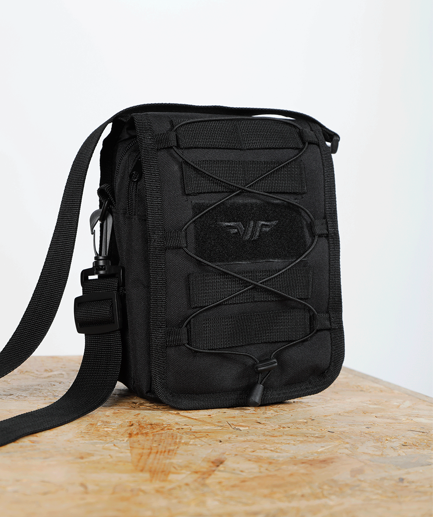 close up view for small, black crossbody shoulder bag with an adjustable strap, front elastic cord, and a subtle logo on the front, displayed against a plain light gray background.