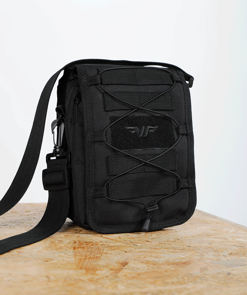  close up view for small, black crossbody shoulder bag with an adjustable strap, front elastic cord, and a subtle logo on the front, displayed against a plain light gray background.